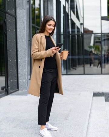 Beautiful young businesswoman wearing autumn coat walking outdoors, holding takeaway coffee cupの写真素材