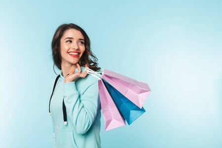 Portrait of a beautiful excited young brunette woman standing isolated over blue background, carrying shopping bagsの写真素材
