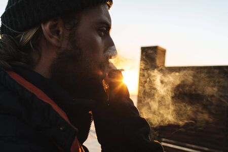 Close up of a man wearing hat standing at the beach during sunset, smoking