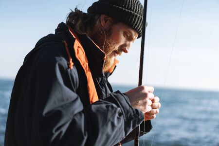 Image of a handsome concentrated young man fisherman wearing coat and hat at the seashore.