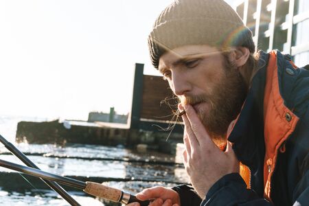 Picture of a handsome young man fisherman wearing coat and hat at the seashore smoking.