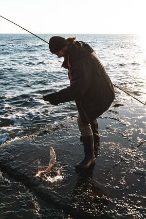 Silhouette of man fisherman wearing coat, holding rod, fishing at the beach