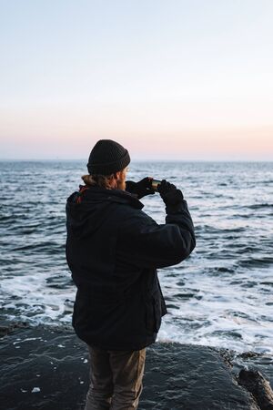 Close up side view of a man silhouette wearing hat standing at the beach during sunset, taking a pictureの写真素材