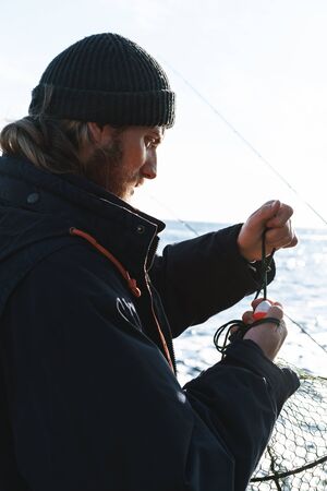 Image of a handsome concentrated young man fisherman wearing coat and hat at the seashore.
