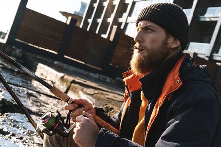 Image of a handsome concentrated young man fisherman wearing coat and hat at the seashore.