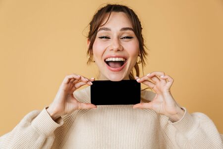 Beautiful cheerful young woman wearing sweater standing isolated over beige background, presenting blank screen mobile phoneの写真素材