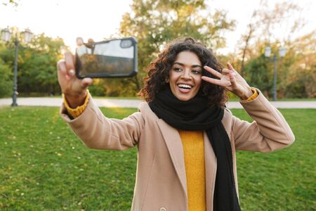 Image of a beautiful young african happy woman walking outdoors in a spring park using mobile phone...