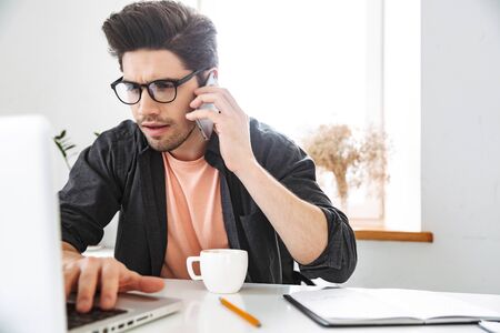 Serious worried handsome man in eyeglasses talking by smartphone and using laptop computer while working by the table at officeの写真素材