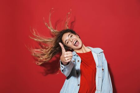 Portrait of a cheerful stylish young woman wearing denim jacket standing isolated over red background, posing, thumbs upの写真素材