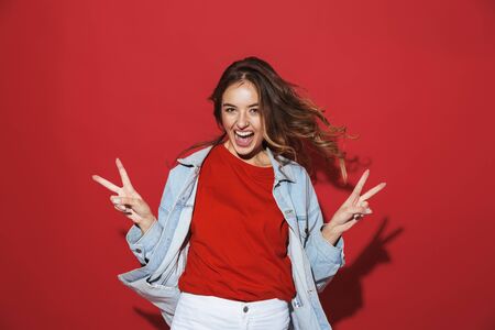 Portrait of a cheerful stylish young woman wearing denim jacket standing isolated over red background, posing, peace gestureの写真素材