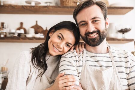 Image of adorable couple man and woman 30s wearing aprons hugging together while cooking in kitchen at homeの写真素材