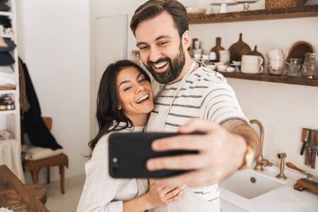 Portrait of beautiful couple man and woman 30s wearing aprons taking selfie photo while cooking...
