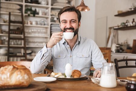 Portrait of joyous european man 30s sitting at table and drinking coffee while having breakfast in kitchen at homeの写真素材