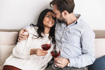 Image of happy brunette couple man and woman drinking wine from glasses together while sitting on couch in bright room at homeの写真素材