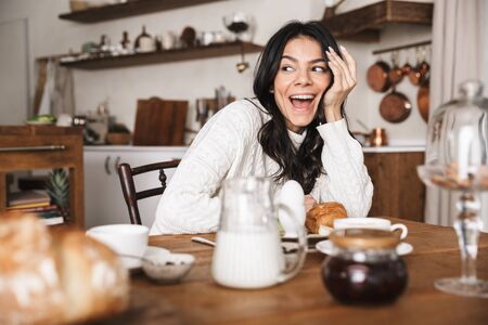 Portrait of happy european woman 30s sitting at table and eating while having breakfast in kitchen at homeの写真素材