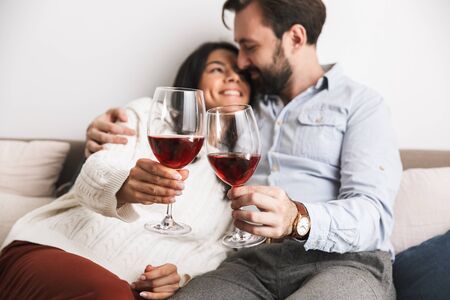 Image of romantic brunette couple man and woman drinking wine from glasses together while sitting on couch in bright room at homeの写真素材