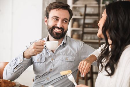 Portrait of caucasian brunette couple man and woman 30s eating together at table while having breakfast in kitchen at homeの写真素材