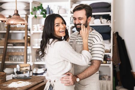 Portrait of young couple man and woman 30s wearing aprons hugging together while cooking in kitchen at homeの写真素材
