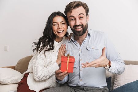 Image of charming couple man and woman holding birthday present box while sitting on sofa in bright room at homeの写真素材