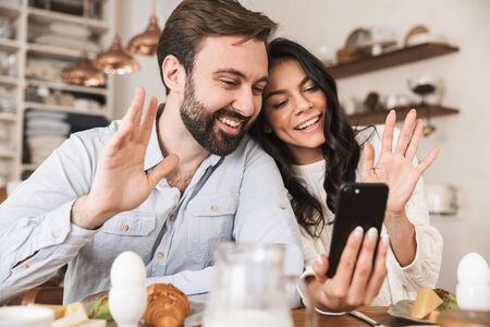 Portrait of adorable european couple man and woman 30s using smartphone while having breakfast in kitchen at homeの写真素材