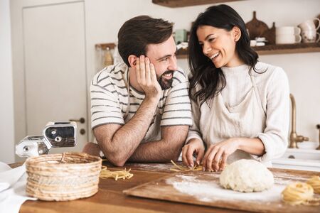 Portrait of positive couple man and woman 30s wearing aprons making homemade pasta of dough while cooking together in kitchen at homeの写真素材