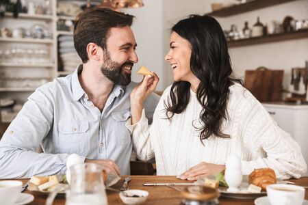 Portrait of attractive brunette couple man and woman 30s eating together at table while having breakfast in kitchen at homeの写真素材