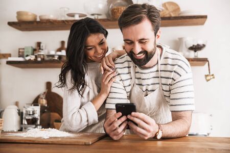 Portrait of joyful couple man and woman 30s wearing aprons reading recipe while cooking pastry with flour and eggs in kitchen at homeの写真素材