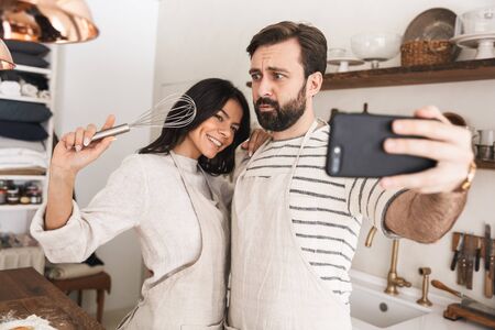 Portrait of optimistic couple man and woman 30s wearing aprons taking selfie photo while cooking pastry with flour and eggs in kitchen at homeの写真素材