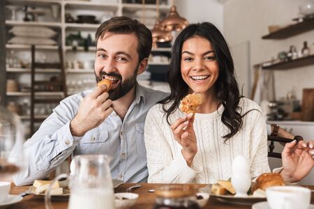Image of young couple man and woman 30s eating together at table while having breakfast in kitchen at homeの写真素材