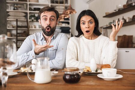 Portrait of surprised couple man and woman 30s eating together at table while having breakfast in kitchen at homeの写真素材