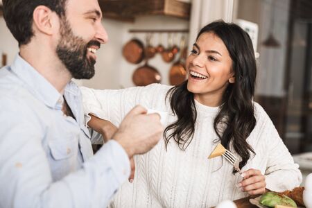 Portrait of positive brunette couple man and woman 30s eating together at table while having breakfast in kitchen at homeの写真素材
