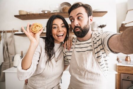 Portrait of funny couple man and woman 30s wearing aprons taking selfie photo while cooking in kitchen at homeの写真素材