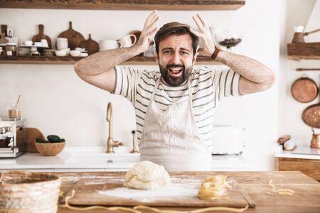 Portrait of nervous brunette man 30s wearing apron cooking and making homemade pasta of dough in kitchen at homeの写真素材