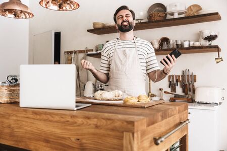 Portrait of joyful brunette man 30s wearing apron using smartphone while cooking and making homemade pasta in kitchen at homeの写真素材