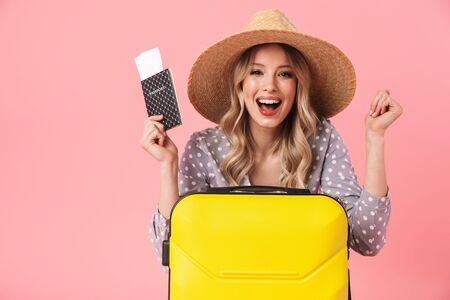 Image of excited young pretty woman tourist posing isolated over pink wall background with suitcase holding passport with tickets make winner gesture.の写真素材