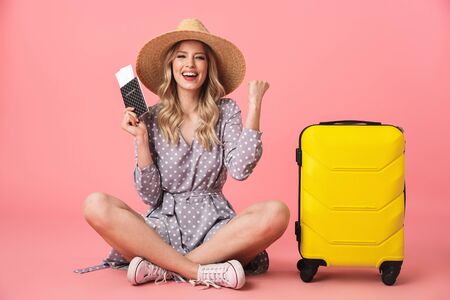 Image of excited young pretty woman tourist posing isolated over pink wall background with suitcase holding passport with tickets make winner gesture.の写真素材