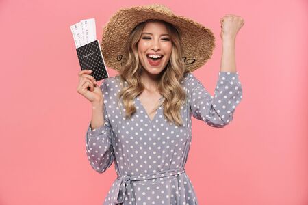 Beautiful young blonde woman wearing summer hat and dress standing isolated over pink background,...