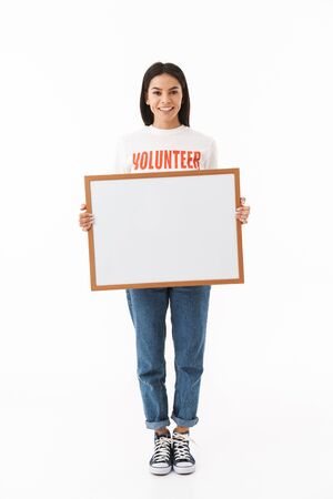 Smiling young girl wearing volunteer t-shirt standing isolated over white background, showing blank boardの写真素材