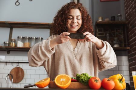 Image of beautiful caucasian woman taking photo of food on smartphone while cooking fresh vegetables salad in kitchen interior at homeの写真素材