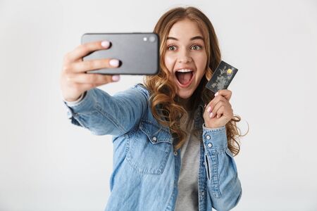 Attractive excited young girl standing isolated over white background, taking a selfie, showing plastic credit cardの写真素材