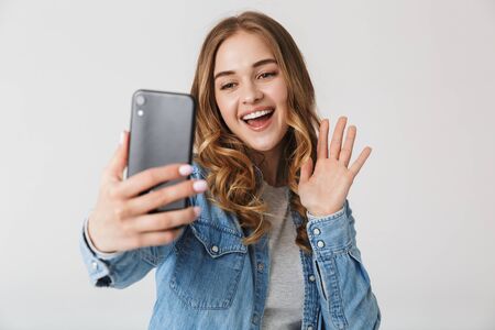 Attractive excited young girl standing isolated over white background, taking a selfie, wavingの写真素材