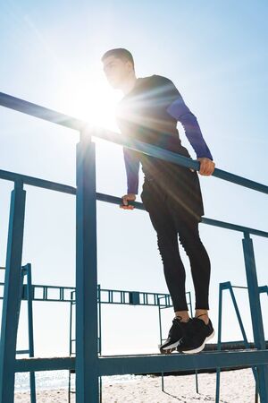 Photo of athletic guy 20s in black tracksuit doing acrobatics on horizontal gymnastic bar during...