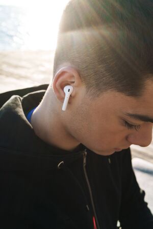 Photo of young sportsman 20s in tracksuit wearing earpods walking by seaside in the morning while...