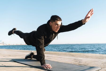 Photo of caucasian sportsman 20s in tracksuit lying on fitness mat and doing plank exercise while...
