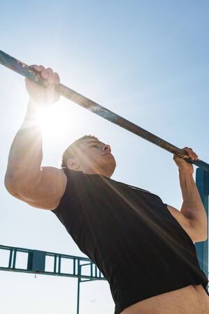 Photo of masculine guy 20s in black tracksuit doing pull ups on horizontal gymnastic bar during...