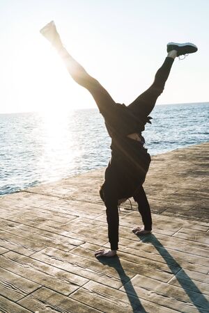 Confident young sportsman wearing jacket warming up at the beach, doing acrobatic exercises