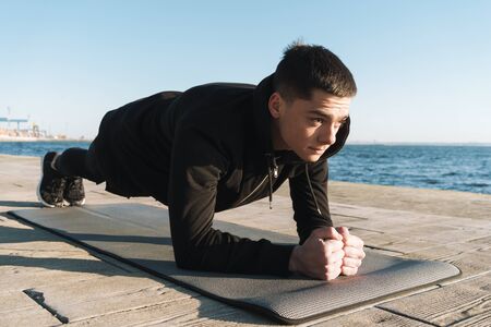 Photo of european sportsman 20s in tracksuit lying on fitness mat and doing plank exercise while...