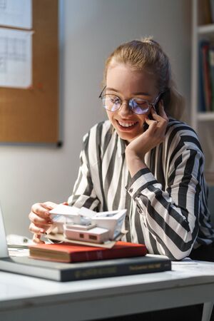 Photo of positive caucasian woman architect wearing glasses holding cellphone and house model while sitting at workplaceの写真素材