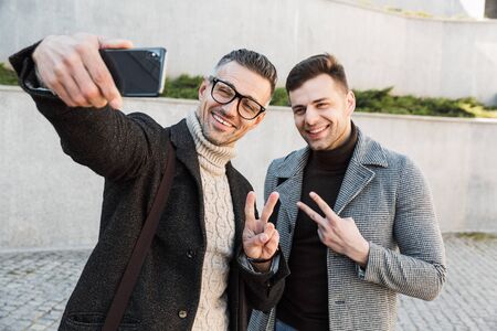 Two handsome men wearing coats spending time outdoors, drinking takeaway coffee, walking, talking,...