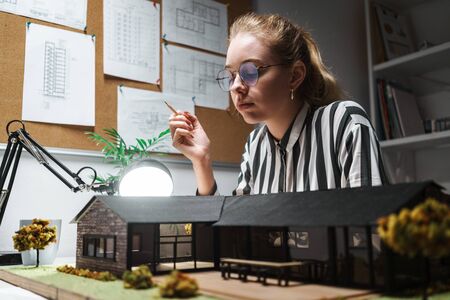 Photo of thoughtful blonde woman architect wearing glasses designing draft with house model and...
